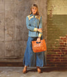 A sustainably sourced woman in a denim shirt and wide leg pants holding a brown leather purse from Bed Stu called the Mista.
