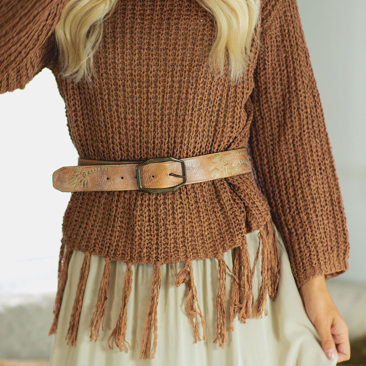 A person wearing the Bed Stu Mohawk brown textured sweater with a fringe hem, paired with a women's distressed light brown leather belt and a cream skirt.