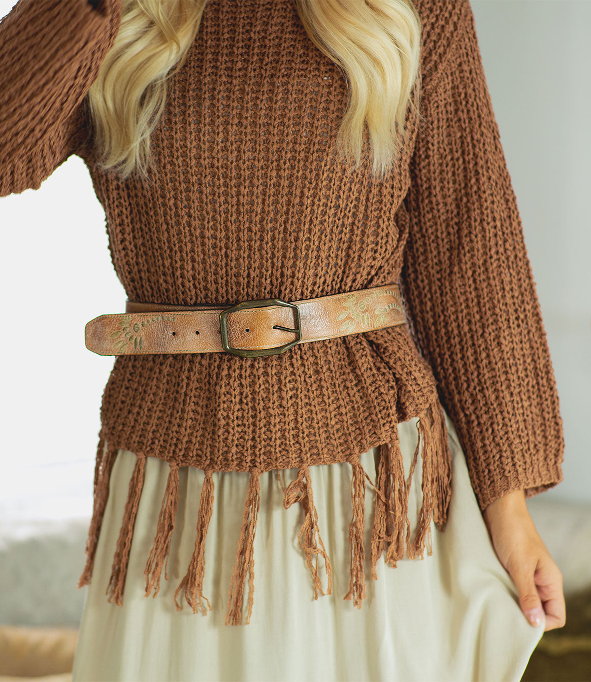 A person wearing the Bed Stu Mohawk brown textured sweater with a fringe hem, paired with a women's distressed light brown leather belt and a cream skirt.