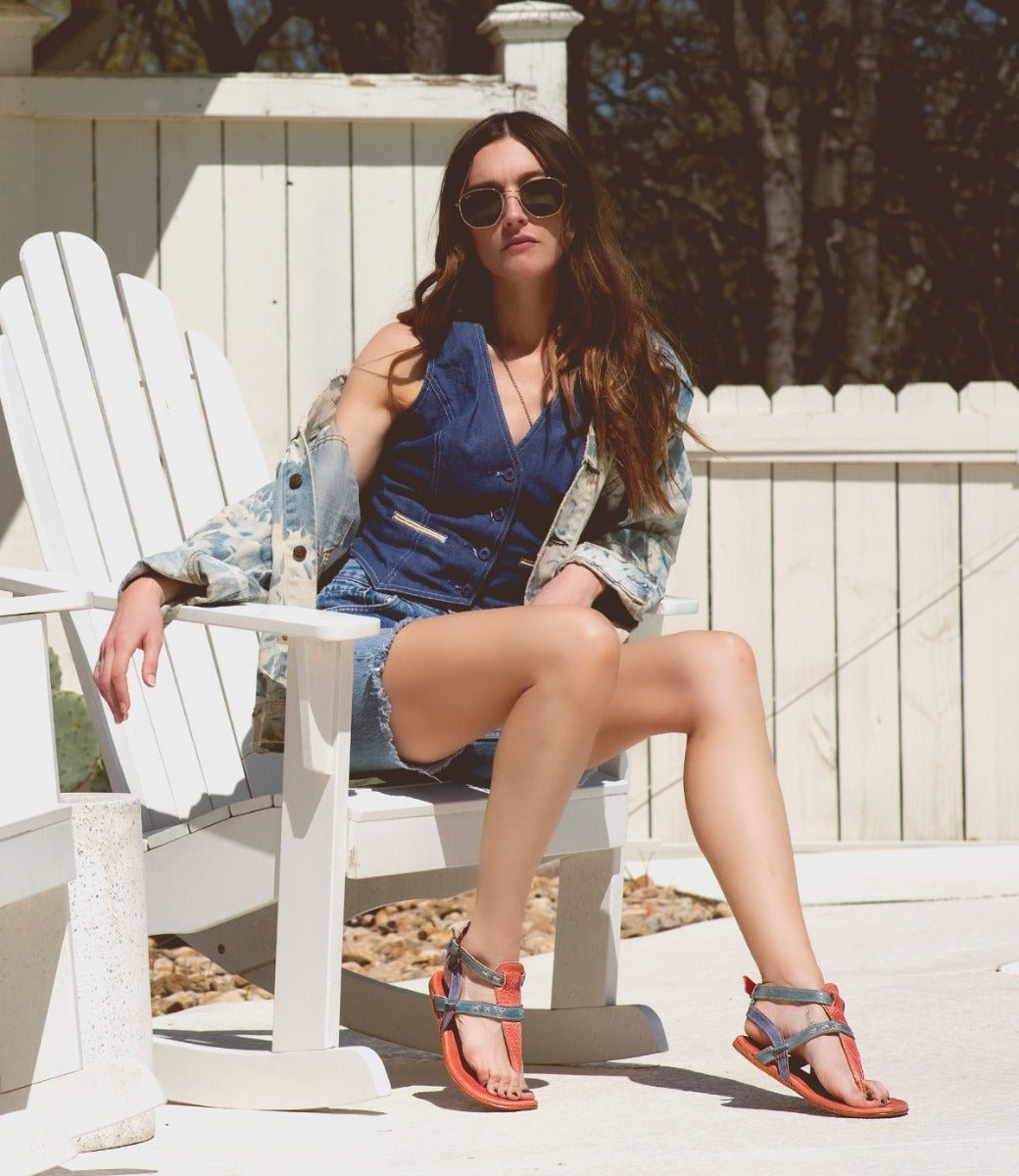 A woman sitting on a white chair wearing Moon sunglasses and a Bed Stu denim jacket.