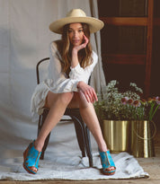 A woman sitting on a chair wearing a blue wedge sandal with a wooden platform by Bed Stu.