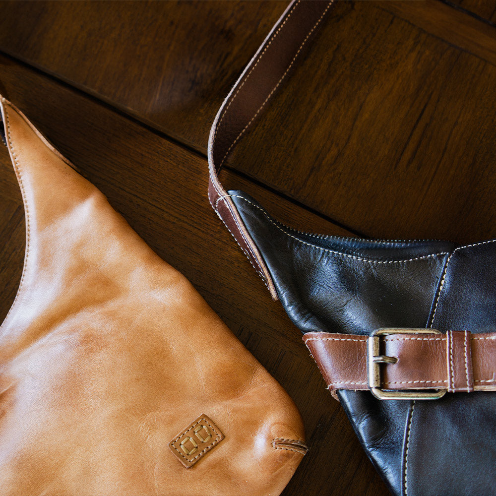 Tan leather bag and black leather shoulder bag with brown trim and a metal buckle arranged on a dark wooden surface.