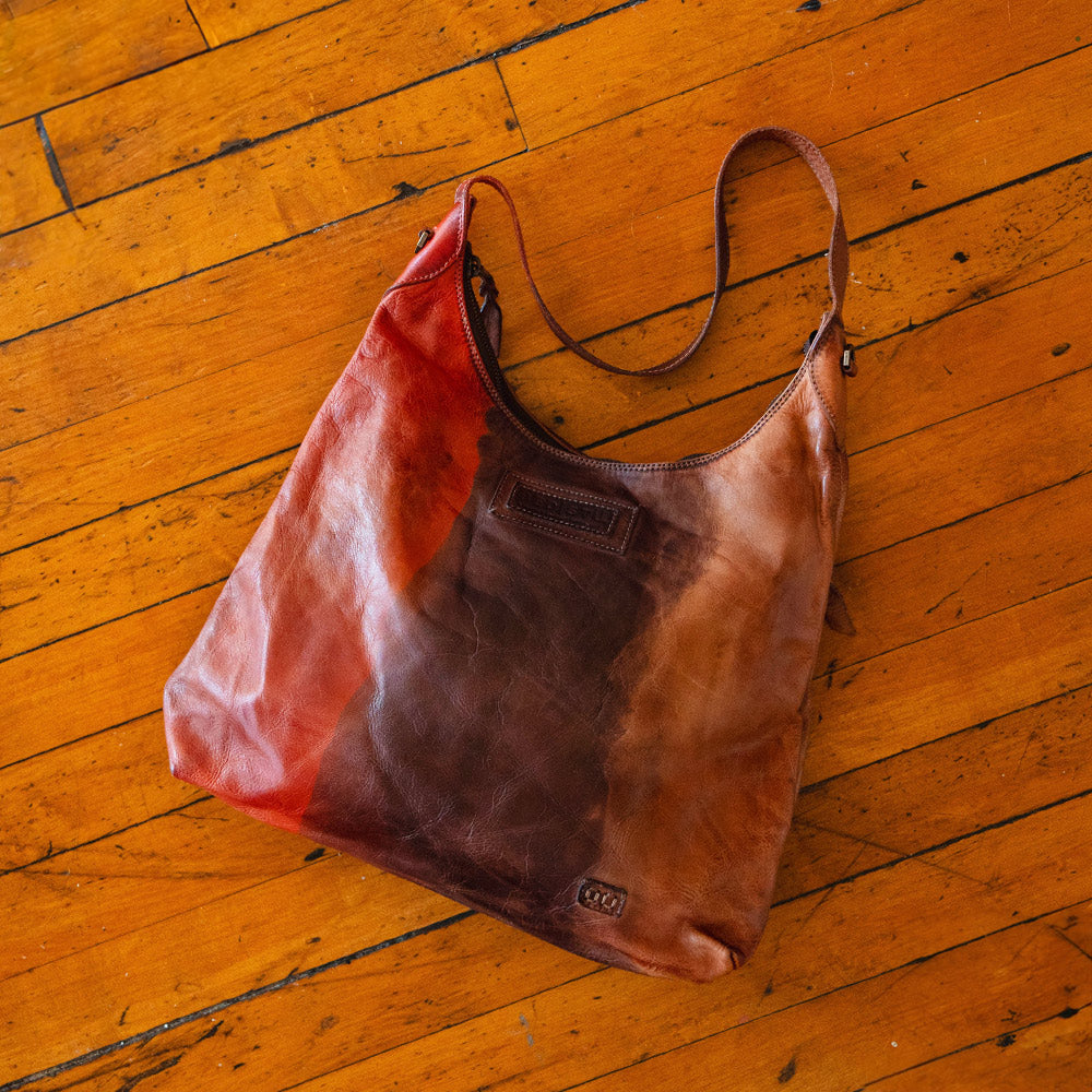 Distressed brown leather shoulder bag with a long strap and multitone reddish-brown finish, resting on a worn wooden floor.