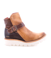 The Bed Stu Nudge II leather ankle boot, in tan and brown, features an adjustable buckle strap, white ridged sole, and convenient side zip, as seen from the side.