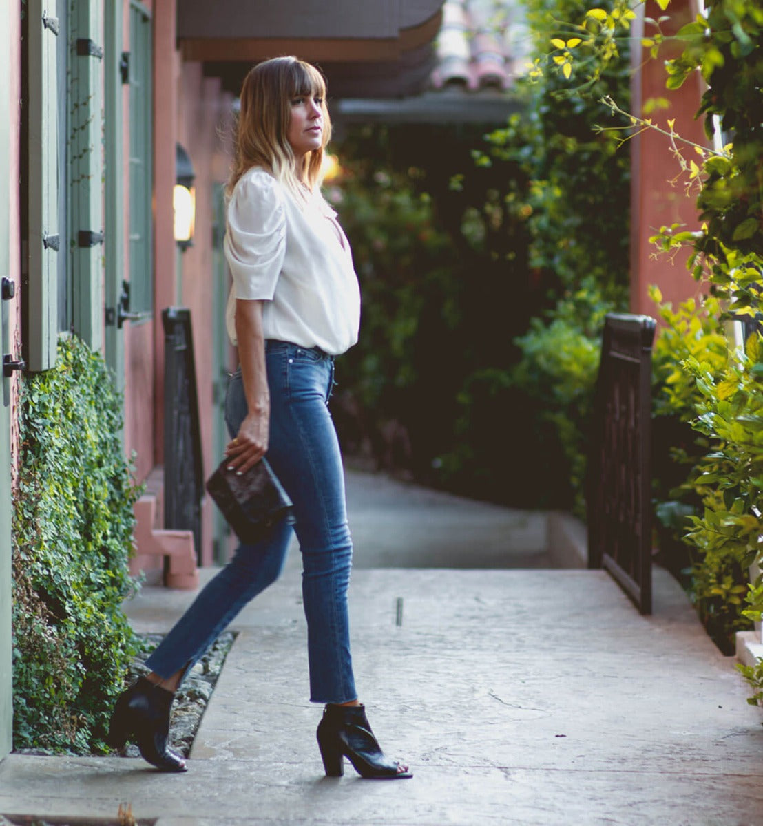 A woman wearing Bed Stu jeans and a Bed Stu white blouse walking down a sidewalk.