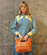 A woman in a denim shirt holding a tan Orchid purse, featuring an adjustable handwoven Bed Stu handbag.