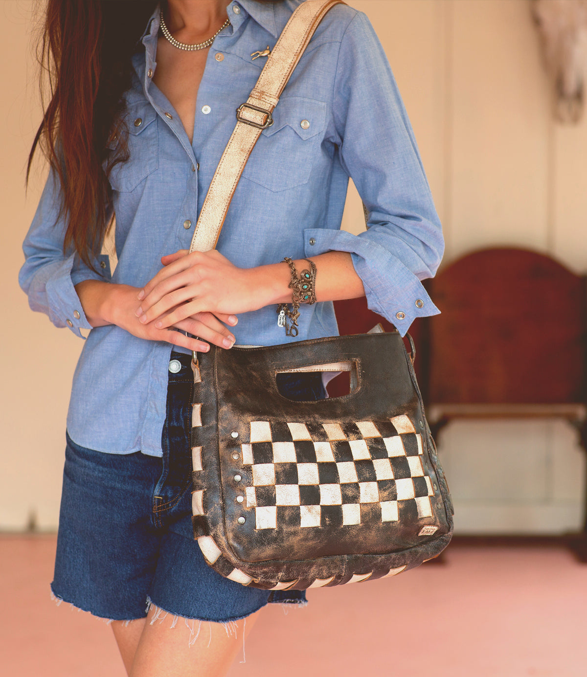 Wearing a blue button-up shirt and denim shorts, a person holds the Bed|Stü Orchid shoulder bag—a black and white checkered accessory featuring a modern, timeless design with an antique metal buckle.