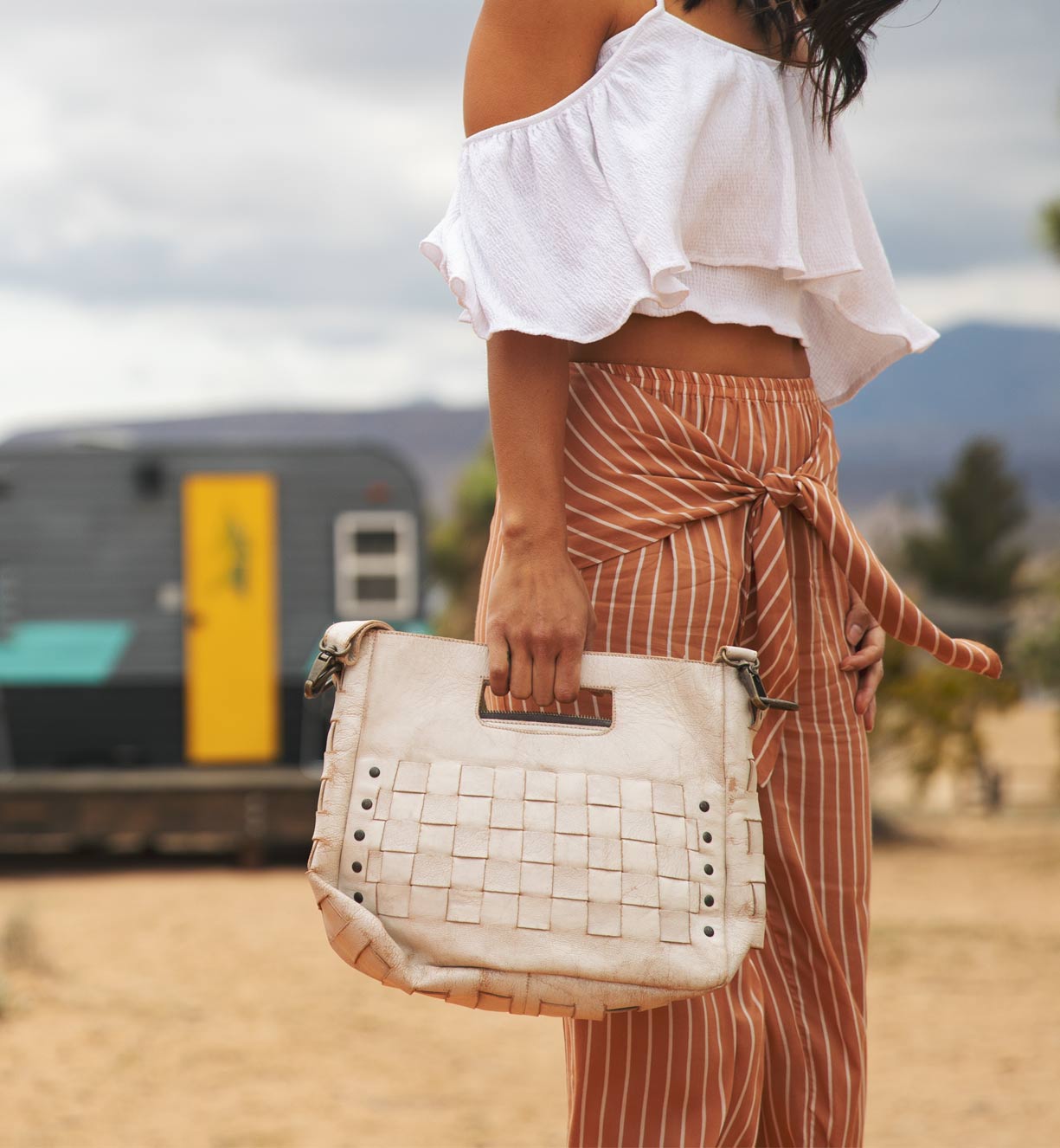 A woman in a white top and striped pants holding a distressed white Bed Stu Orchid bag.