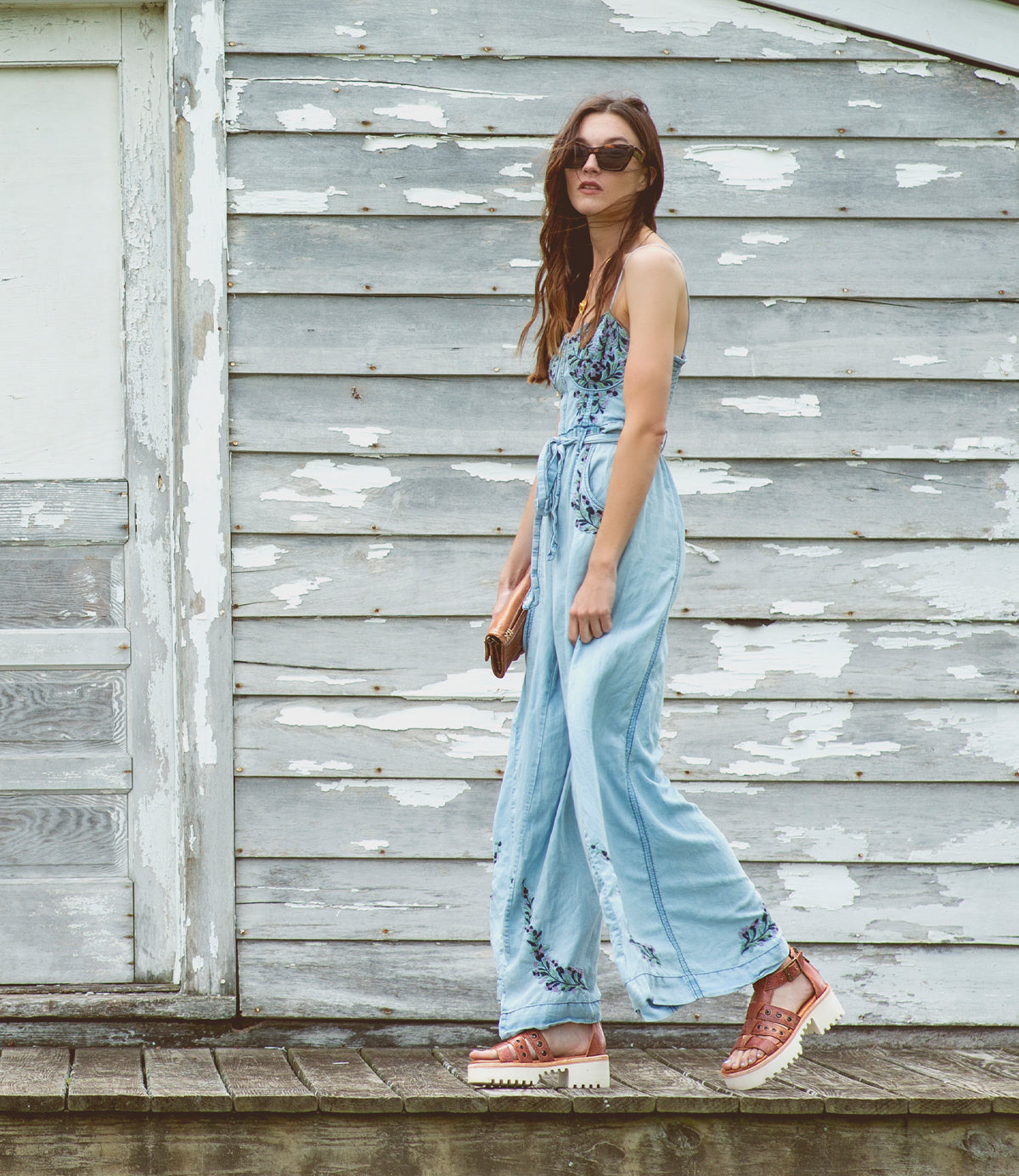 A woman wearing a jumpsuit and the Pacifica sandals from Bed Stu on a wooden deck.