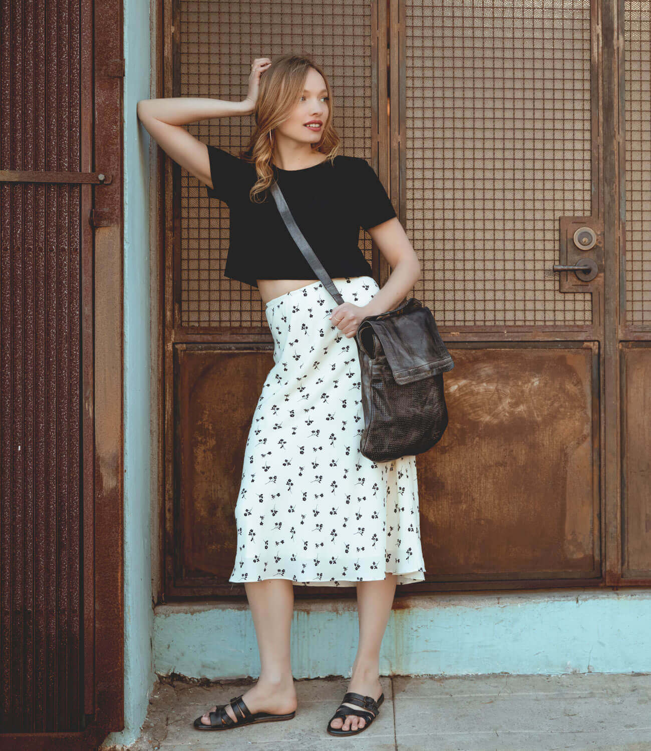 A woman leaning against a wall wearing a black crop top and Bed Stu's Patsy floral midi skirt.