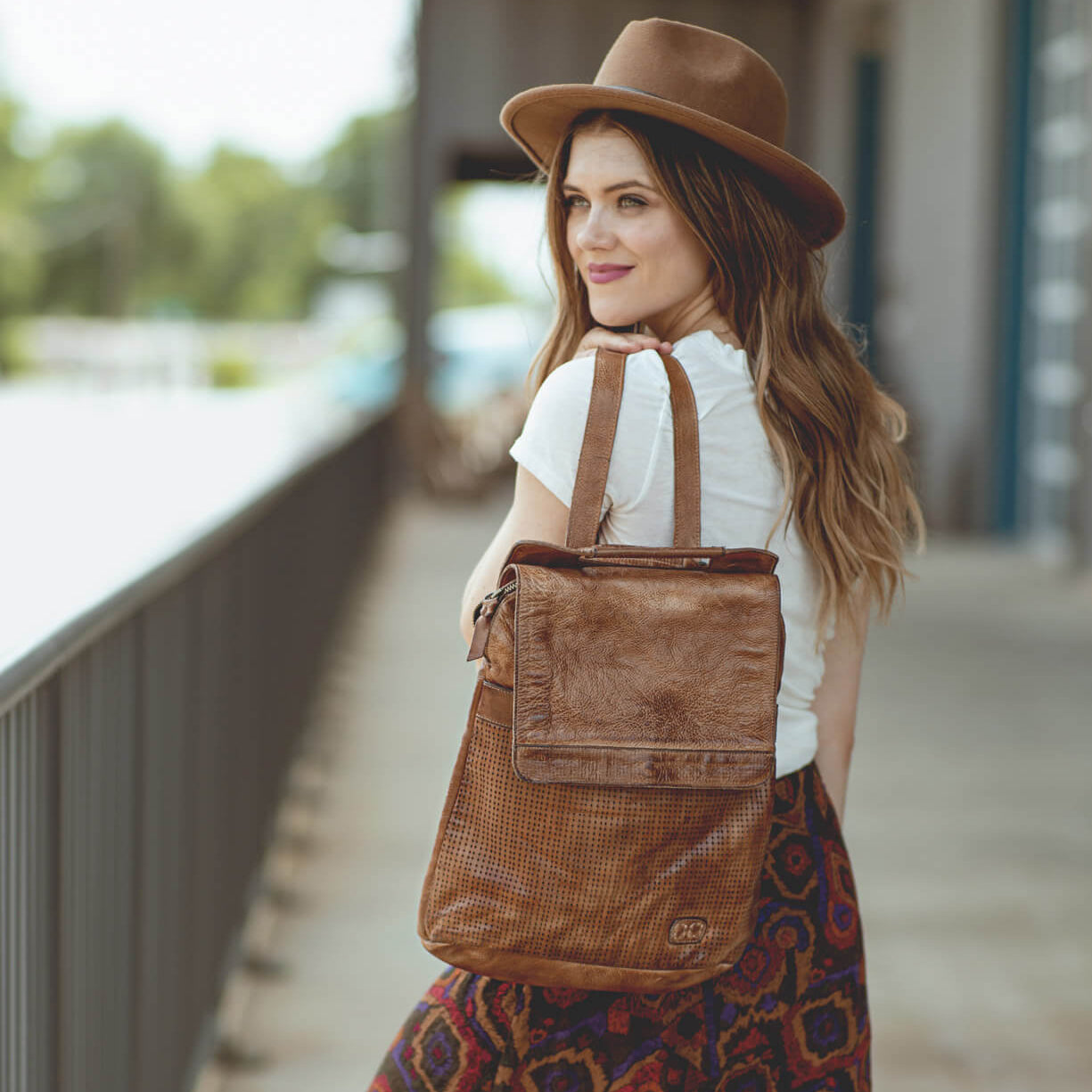 A woman wearing a hat and skirt is holding a Bed Stu Patsy brown leather bag.