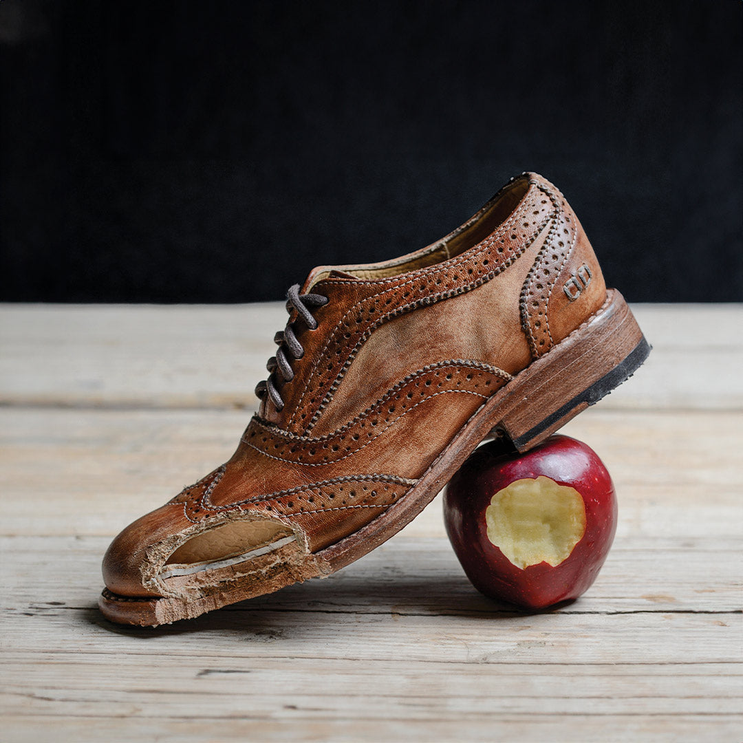 A brown leather brogue shoe with a hole in the toe resting on a bitten red apple.