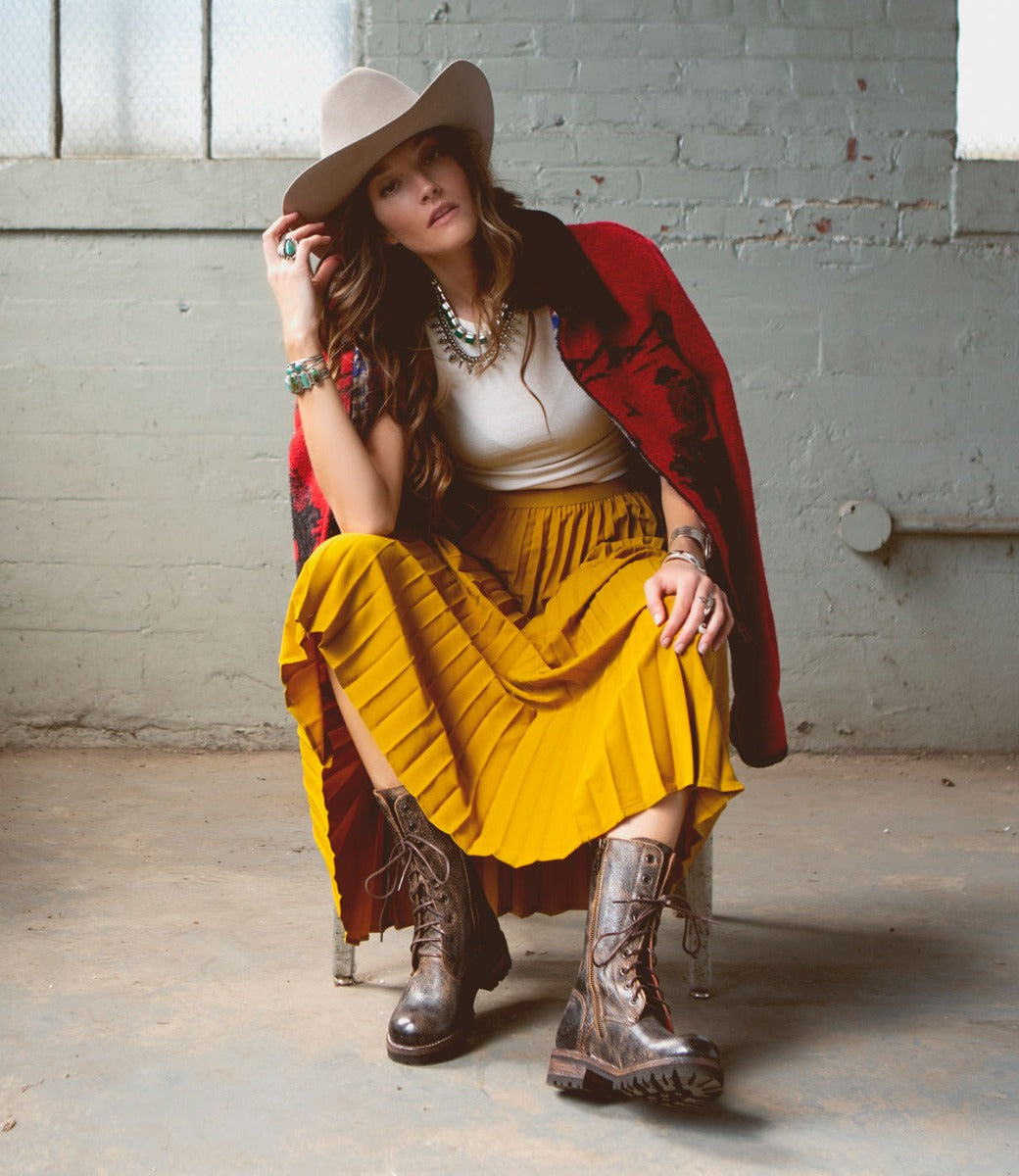 A woman in a yellow skirt and hat sitting on a chair wearing Posh boots from Bed Stu.