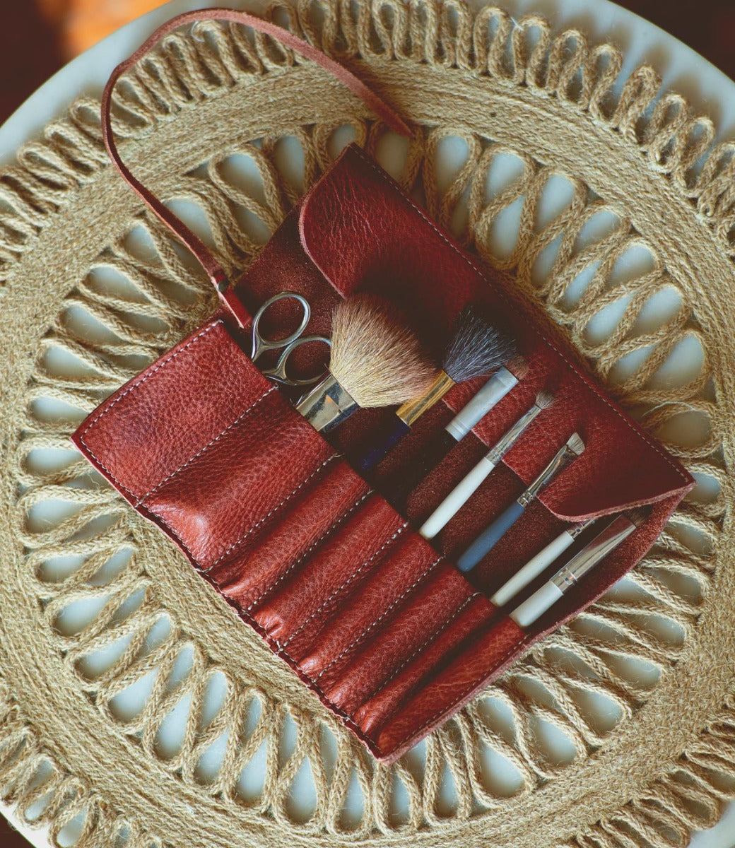 A red Prepped Leather Wrap from Bed Stu, containing various makeup brushes and a pair of small scissors, is open on a woven circular mat.