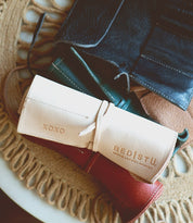 A close-up of several leather pencil rolls in various colors, laid on a woven placemat. One roll in the foreground from Bed Stu features the text "XOXO" and "BEDIŠTU" embossed on it—perfect for organizing artist tools.