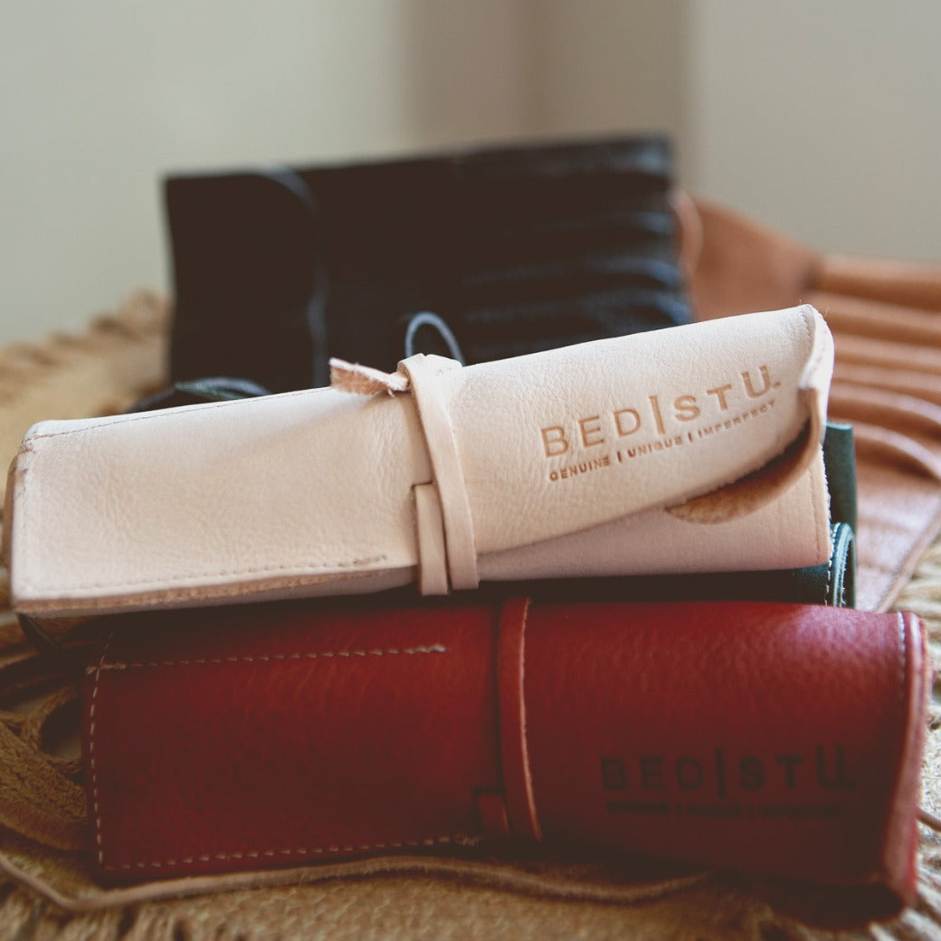 Close-up of rolled-up Prepped Leather Wrap in various colors, including white, black, and red, embossed with the brand name "Bed Stu." These artist tools are arranged on a textured fabric surface, showcasing their craftsmanship from the Leather Shoppe.