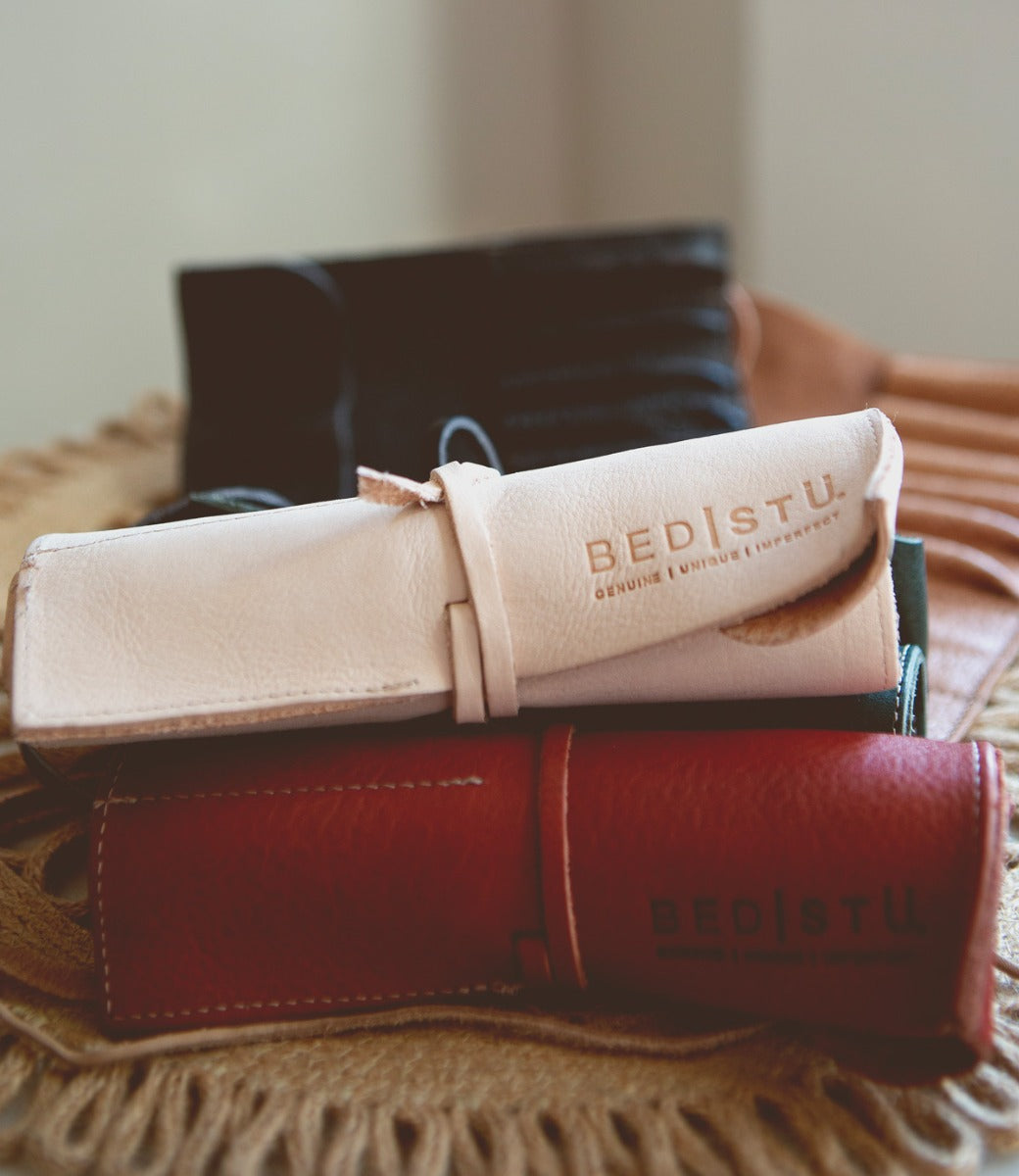Close-up of rolled-up Prepped Leather Wrap in various colors, including white, black, and red, embossed with the brand name "Bed Stu." These artist tools are arranged on a textured fabric surface, showcasing their craftsmanship from the Leather Shoppe.