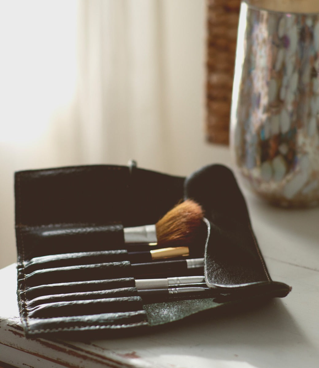 A set of five makeup brushes in a Prepped Leather Wrap from Bed Stu lies open on a wooden table beside a decorative vase.