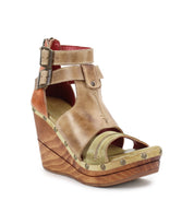 The "Princess" sandal by Bed Stu is a tan leather wedge featuring a sustainable wooden platform, open toe, woven strappy uppers, and a double ankle buckle system with metal studs.