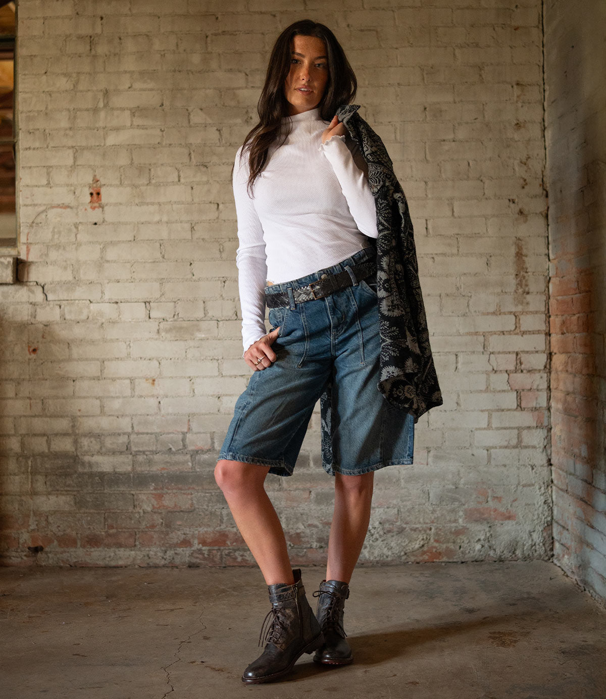 A woman stands indoors against a brick wall, wearing Bed Stu's Proem rustic casual belt with a vintage metal buckle, paired with a white long-sleeve top, denim Bermuda shorts, ankle boots, and holding a patterned jacket over her shoulder.