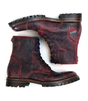 A pair of red and black leather combat style boots with rugged Vibram outsoles from Bed Stu's Protege Trek collection, shown from the side. One boot is placed horizontally, while the other stands perpendicular on top of it, showcasing their sturdy design.