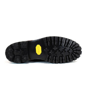 The image shows the underside of a black rubber-soled Bed Stu Protege Trek combat style boot with deep treads and a yellow hexagonal Vibram outsole logo in the center.