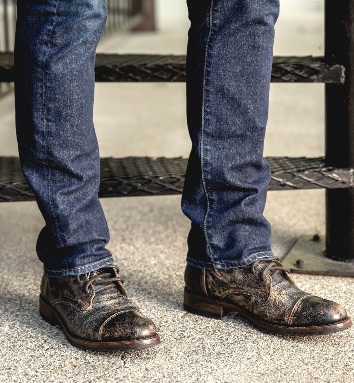 A person wearing dark blue jeans and Bed|Stü's Protege distressed brown leather lace-up boots, styled like a classic Goodyear welt combat boot, stands on a concrete surface next to a metal stair.