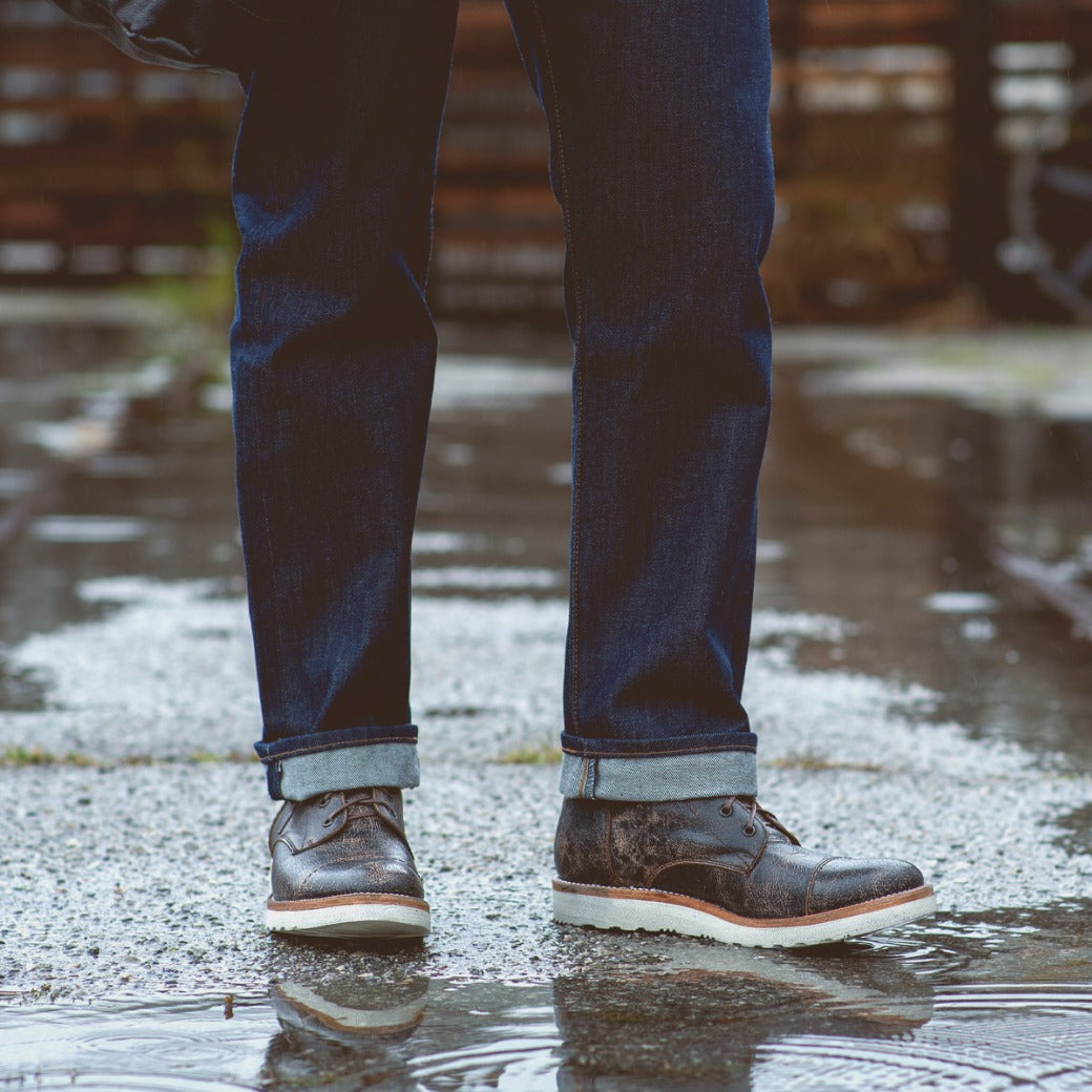 A person wearing dark jeans and Bed Stu Protege Light men's boots with cushioned leather insoles, standing in a puddle on a rainy day.