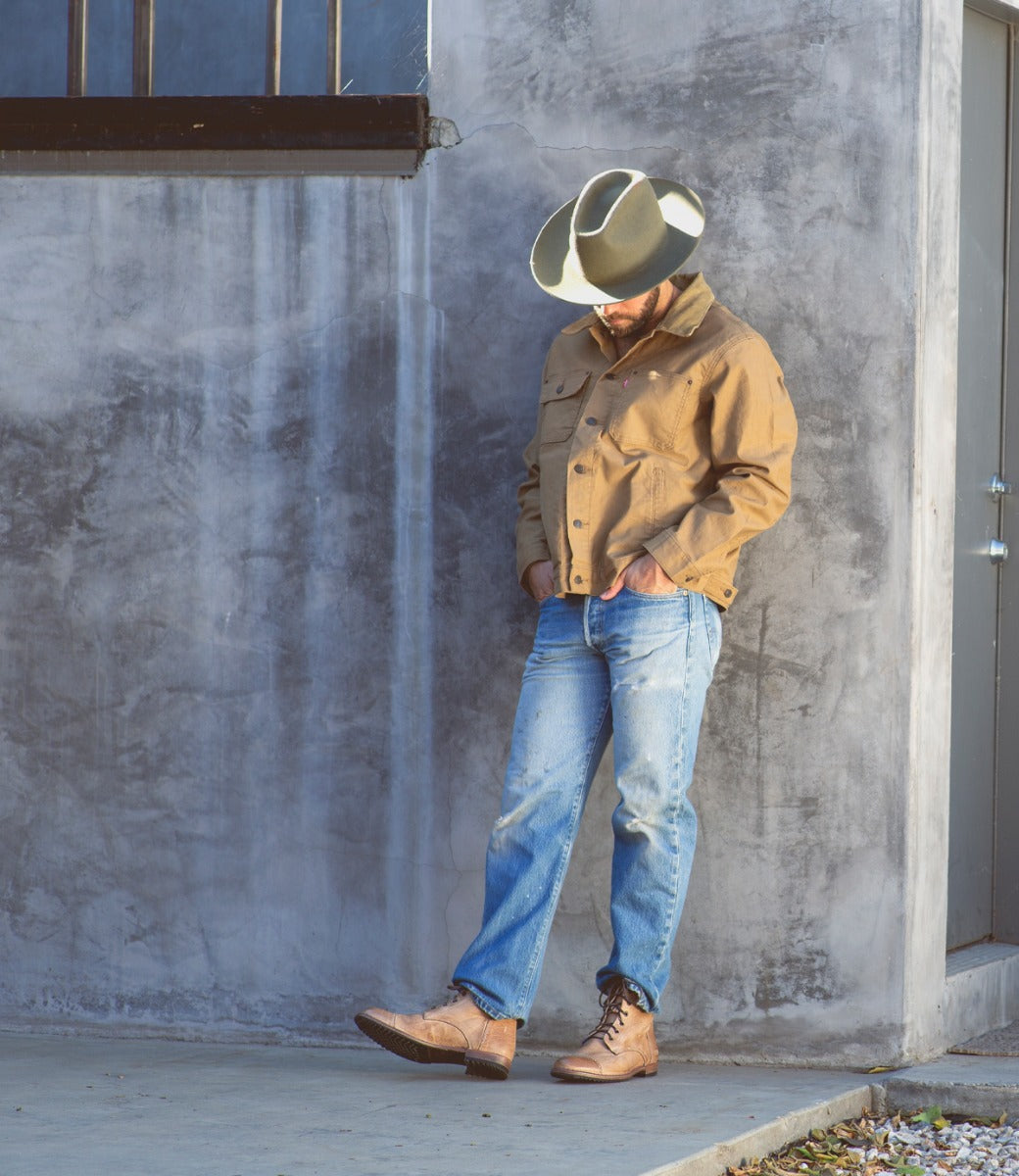 A man in a cowboy hat leaning against a wall, wearing tan Protege boots.