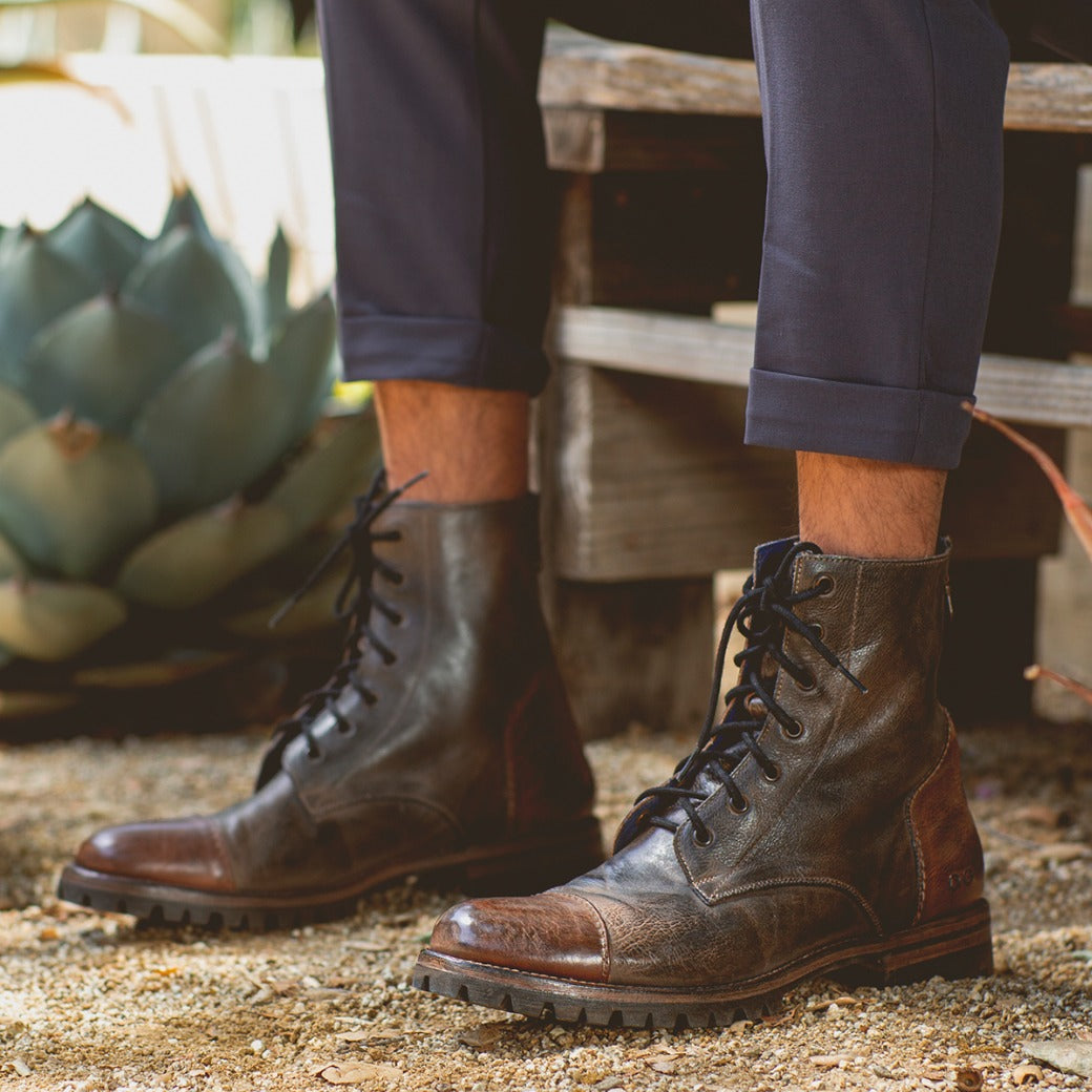 A person wearing Bed Stu Protege Trek boots and navy pants sits on a wooden bench, surrounded by plants and gravel.