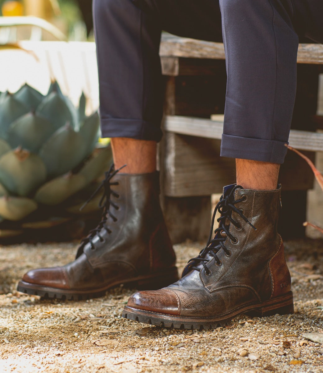 A person wearing Bed Stu Protege Trek boots and navy pants sits on a wooden bench, surrounded by plants and gravel.