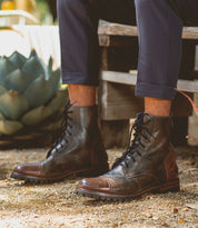 A person wearing Bed Stu Protege Trek boots and navy pants sits on a wooden bench, surrounded by plants and gravel.