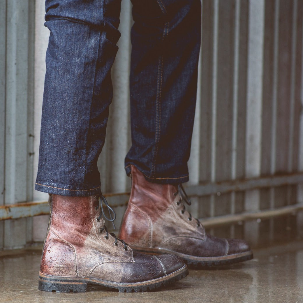 A person wearing rolled-up Protege Trek jeans and Bed Stu combat style boots stands on a wet surface near a corrugated metal wall.