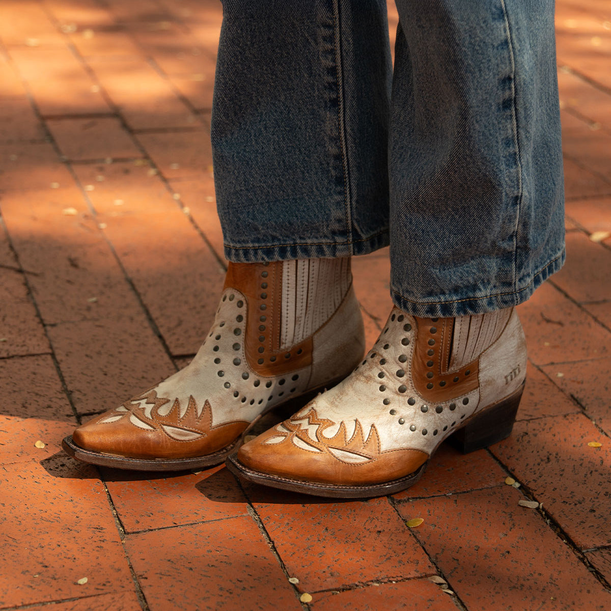 A person wearing blue jeans and Bed Stu’s Raise brown Western boots with decorative patterns and a snip toe, standing on a sunlit brick pavement.
