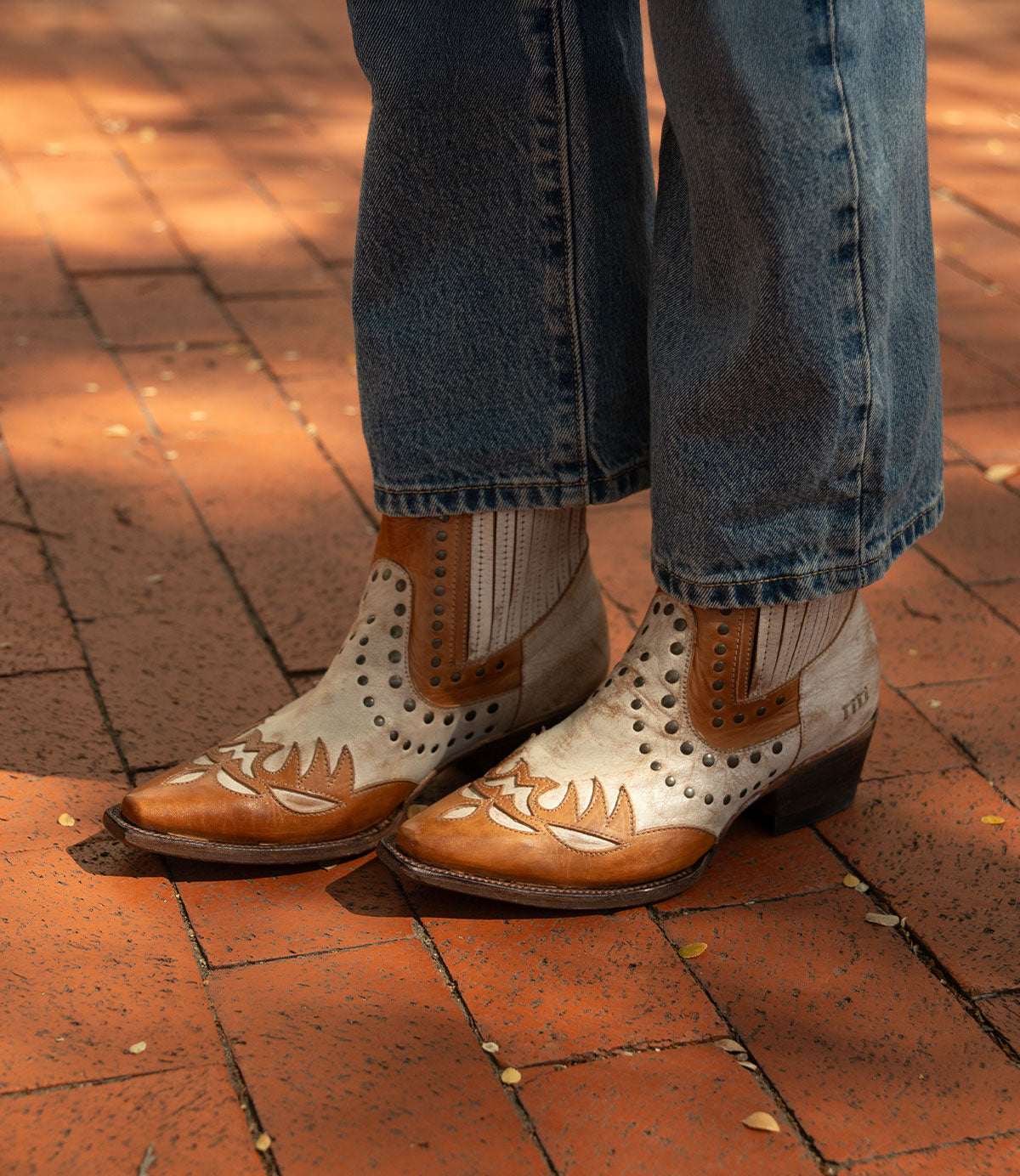 A person wearing blue jeans and Bed Stu’s Raise brown Western boots with decorative patterns and a snip toe, standing on a sunlit brick pavement.