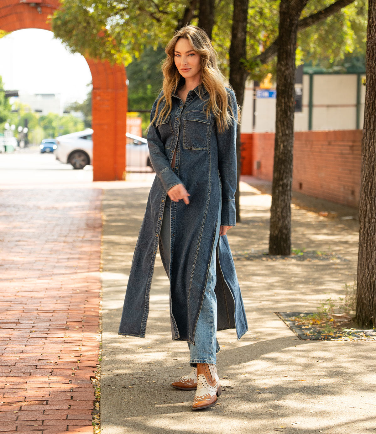 A woman stands on a sunlit sidewalk in jeans, a long denim coat, and Bed Stu's Raise Western boots with a snip toe, with trees and a brick walkway behind her.