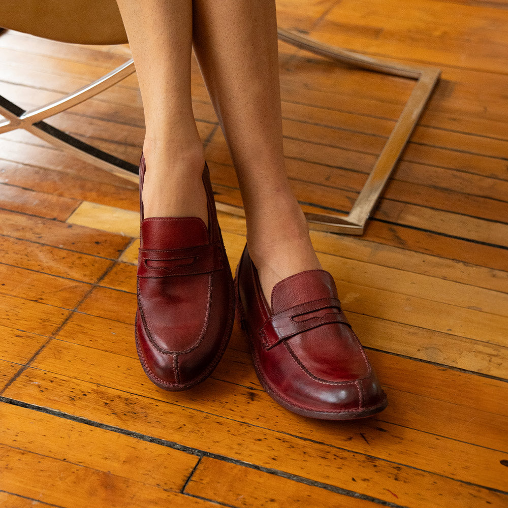 Burgundy leather penny loafers with moc-toe stitching, worn while standing on a warm-toned wooden floor.