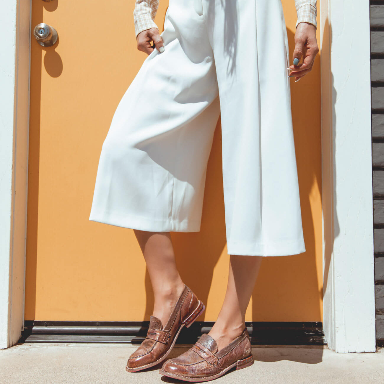 A woman is standing in front of a yellow door wearing white pants and Bed Stu Reina shoes.