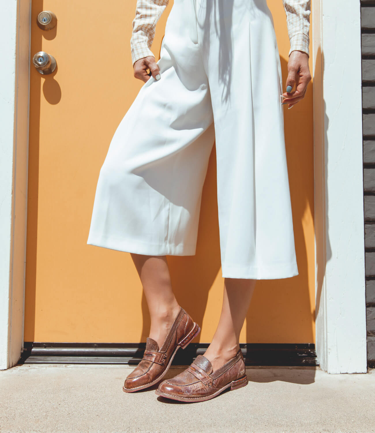 A woman is standing in front of a yellow door wearing white pants and Bed Stu Reina shoes.