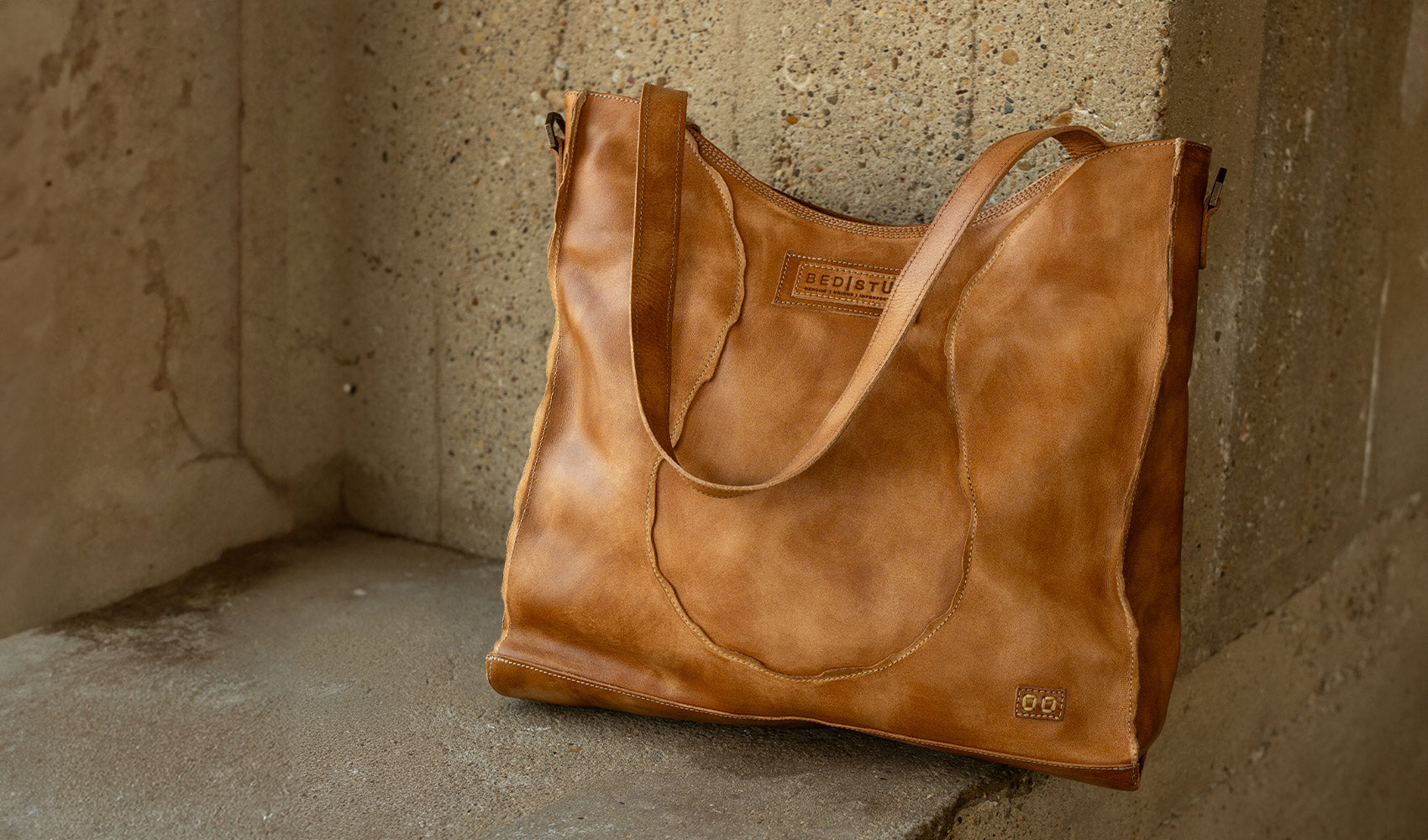 Large tan leather tote bag with two shoulder straps, stitched panel detailing, and a visible BED|STU label, resting against a concrete wall.