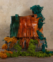 The Bed Stu Rimes handcrafted leather tote with brown and green fringe rests on a metal crate, surrounded by dried green and yellow moss, set against a textured beige wall.