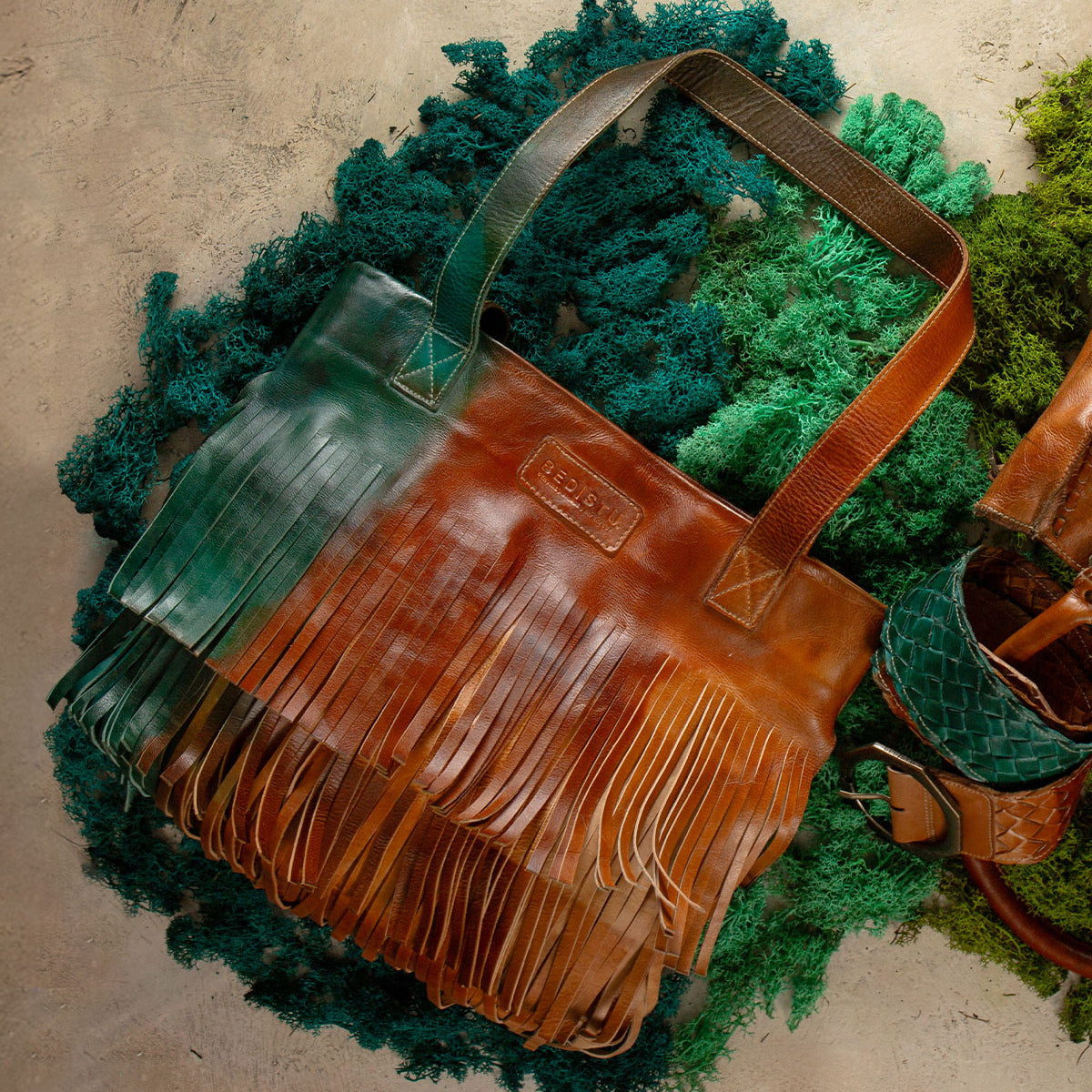 The Rimes tote by Bed Stu features handcrafted leather with green and brown ombre hues and fringe accents, shown resting on a textured green and beige surface.