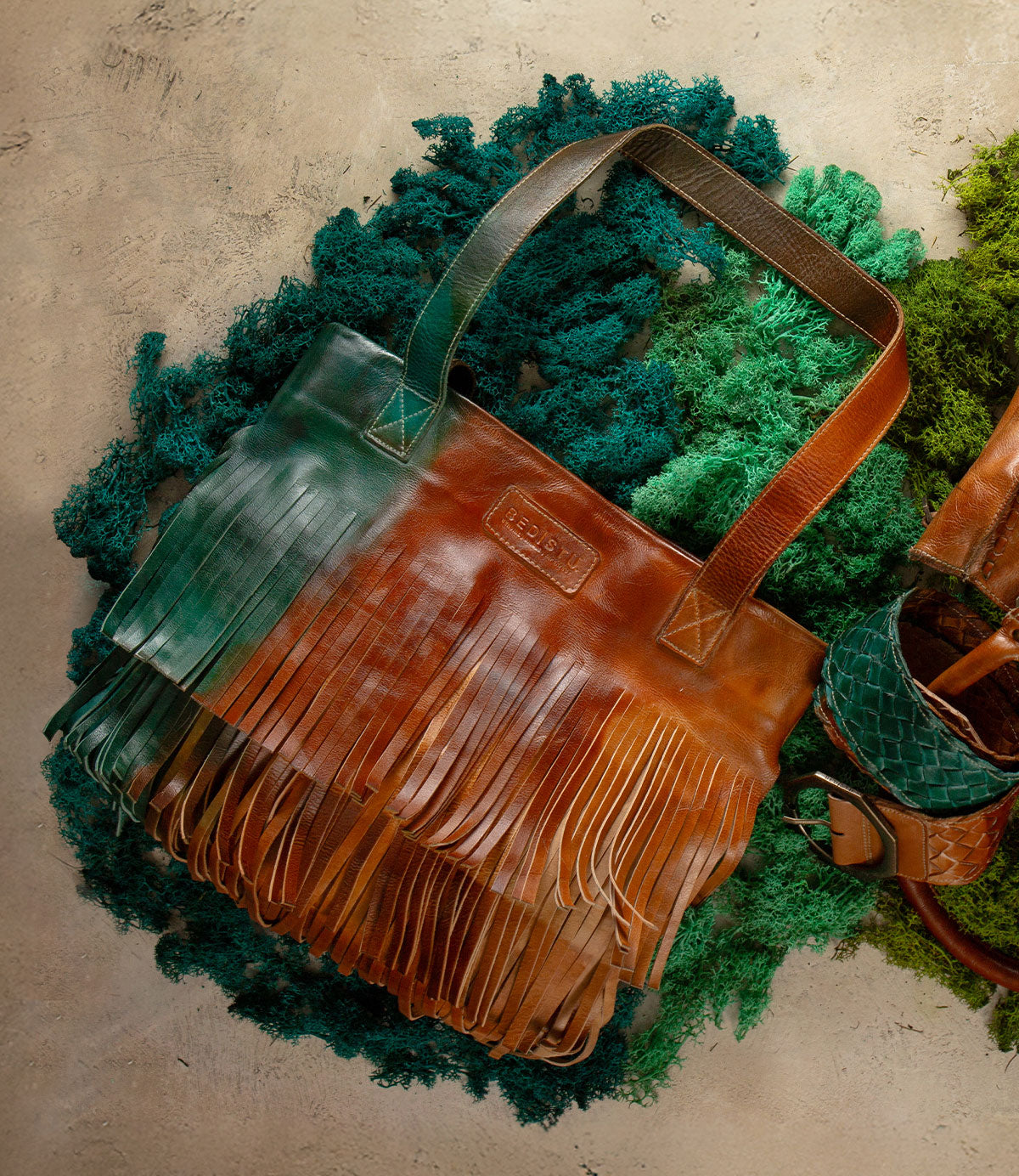 The Rimes tote by Bed Stu features handcrafted leather with green and brown ombre hues and fringe accents, shown resting on a textured green and beige surface.