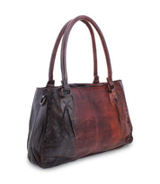 Introducing the Rockababy by Bed Stu: a chic two-toned leather handbag with dual handles, featuring visible stitching and a distressed finish. This rectangular bag is adorned with decorative tassel accents and metal studs along the edges. For added convenience, it also comes with versatile crossbody straps.