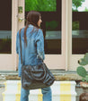 A woman carrying a Rockaway bag on the steps of a house. (Brand: Bed Stu)