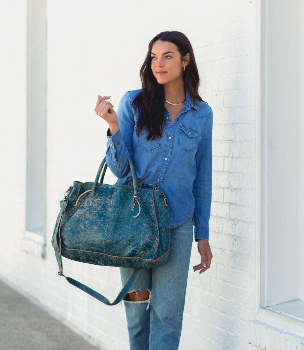 A woman in a blue shirt and jeans holding a Bed Stu Rockaway tote bag.