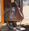 A woman is holding a Rockaway bag in front of a camper. The bag is made by Bed Stu.