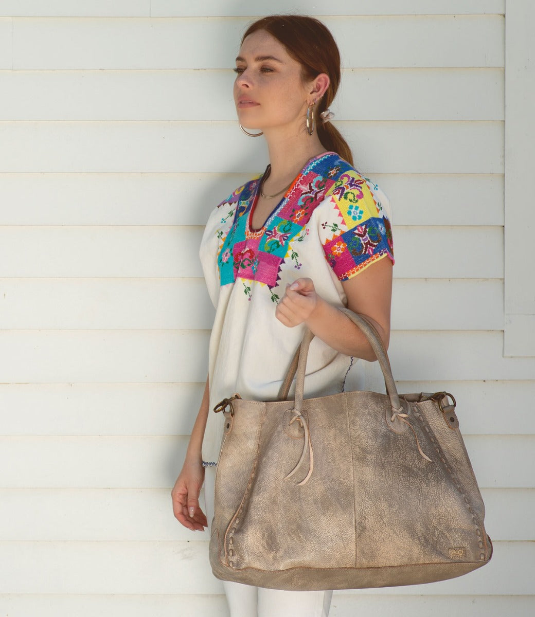 A woman wearing a colorful blouse and white shorts holding a Bed Stu Rockaway tote bag.