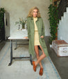 A woman in a green Bed Stu trench coat posing in front of a table.