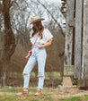 A woman wearing Bed Stu ripped jeans and a Bed Stu cowboy hat in front of a barn.